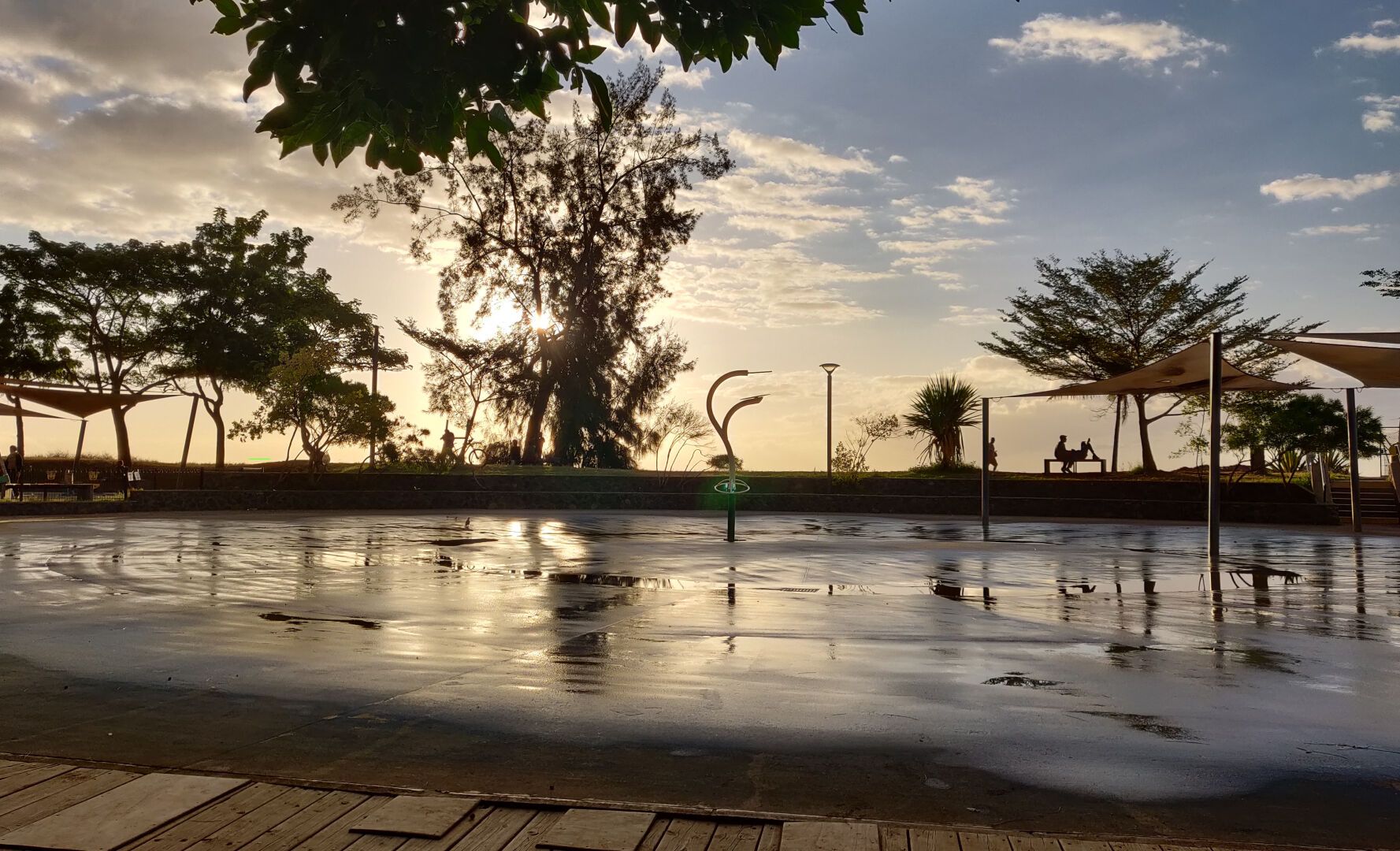 Coucher de soleil sur le front de mer de Saint-Paul, à la Réunion.
On peut voir les arbres et les structures d'une aire de jeux à contre jour avec le soleil qui se couche derrière l'un des arbres, générant des reflets dorés dans les flaques créées par les jets d'eau qui ont fait le bonheur des enfants tout l'après-midi.